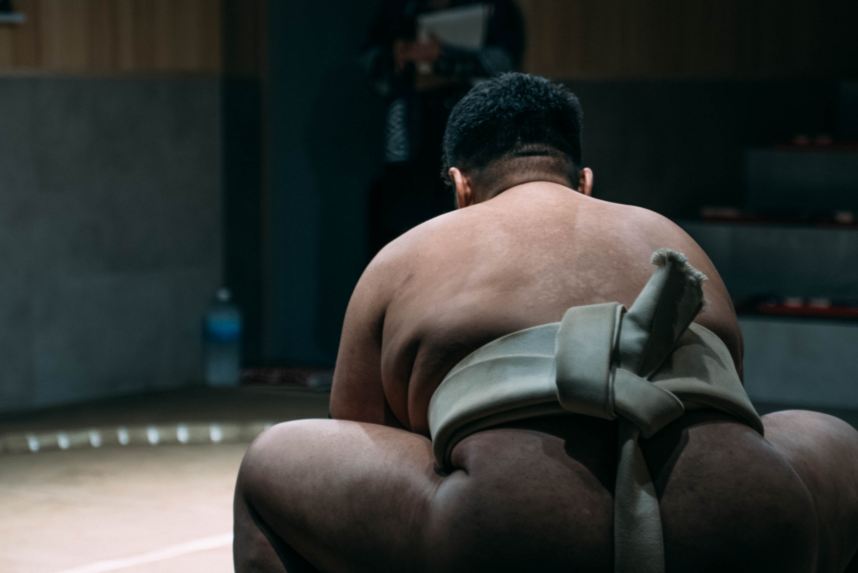 A sumo wrestler crouched low in the ring, seen from behind under dramatic lighting